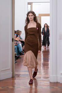Model walks a runway in a brown long-sleeve midi dress with a beige sheer bubble hem and burgundy loafers.