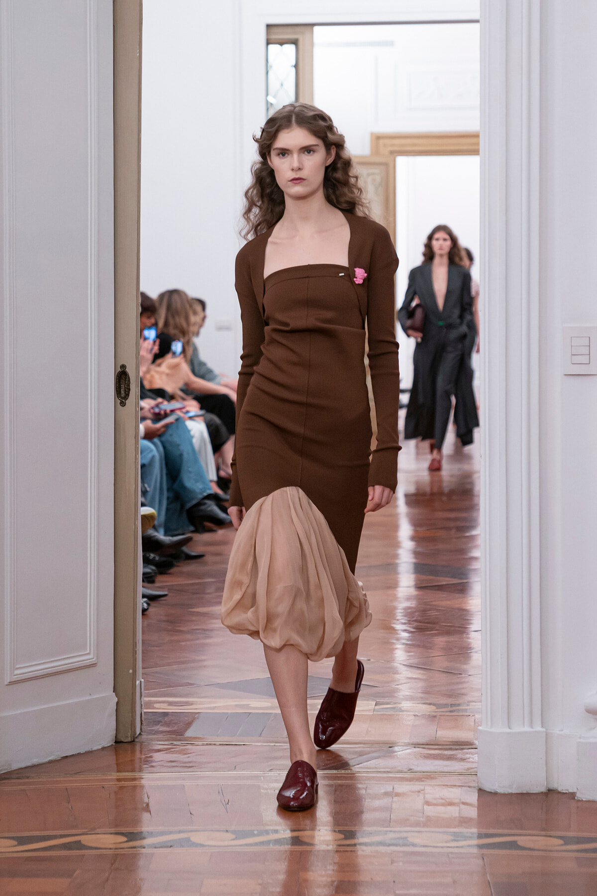 Model walks a runway in a brown long-sleeve midi dress with a beige sheer bubble hem and burgundy loafers.