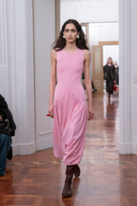 Model walking a fashion show runway in a sleeveless pink dress with a draped skirt and brown knee-high boots, in a bright interior setting.