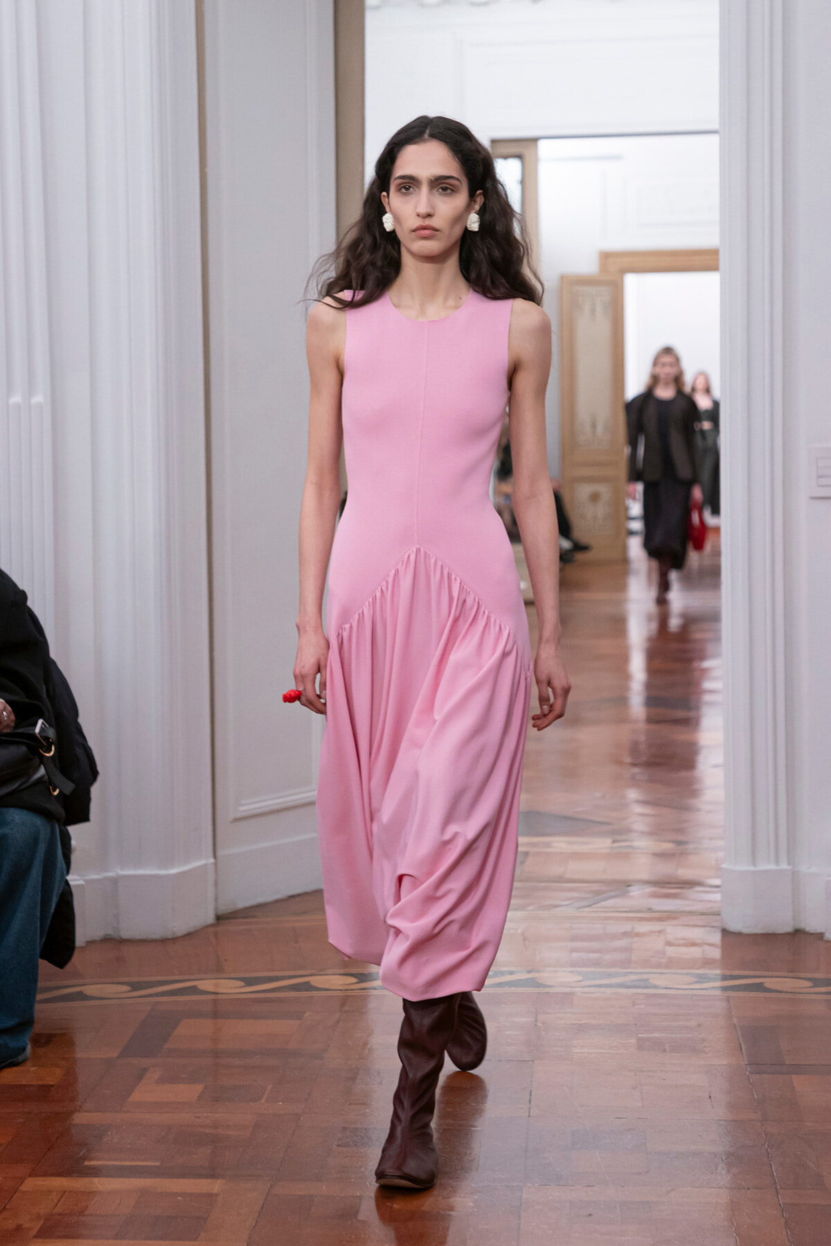 Model walking a fashion show runway in a sleeveless pink dress with a draped skirt and brown knee-high boots, in a bright interior setting.