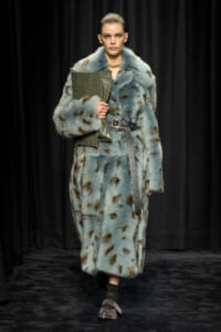 Model walks a runway in a light blue spotted fur coat, belted at the waist, carrying a green textured clutch.