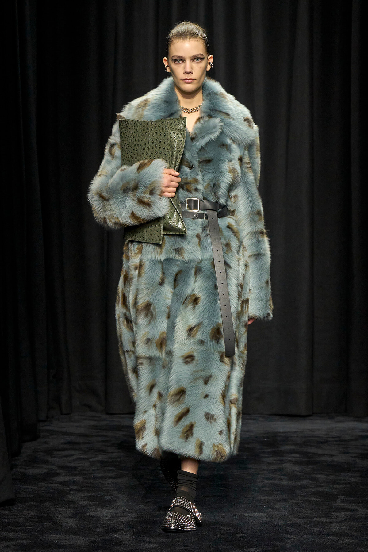 Model walks a runway in a light blue spotted fur coat, belted at the waist, carrying a green textured clutch.