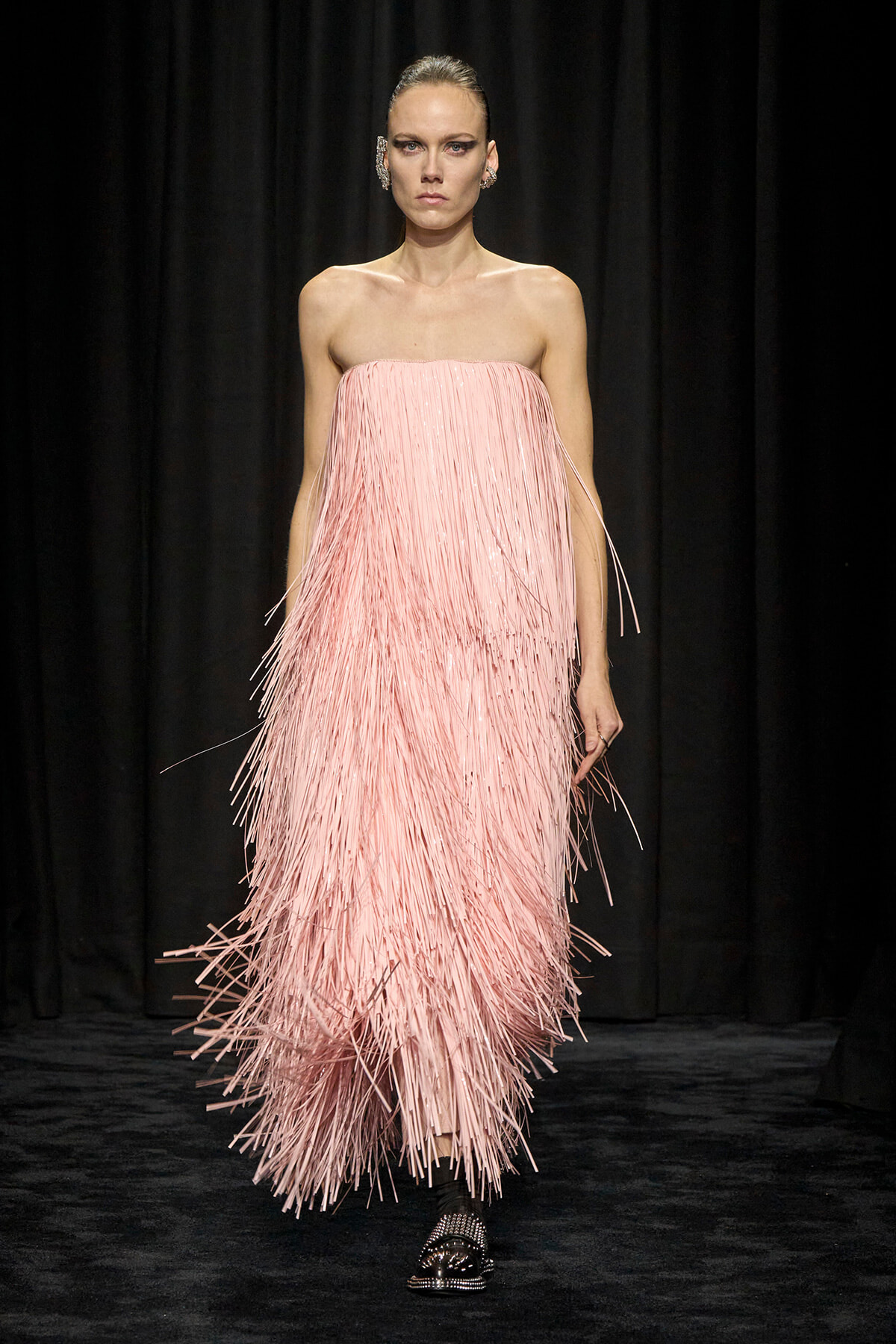 Model walking a runway in a strapless pink fringe gown that sways with movement; black backdrop.