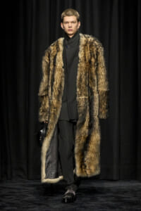 Model on a runway wearing a long fur coat over a black outfit, carrying a black bag, against dark curtains.