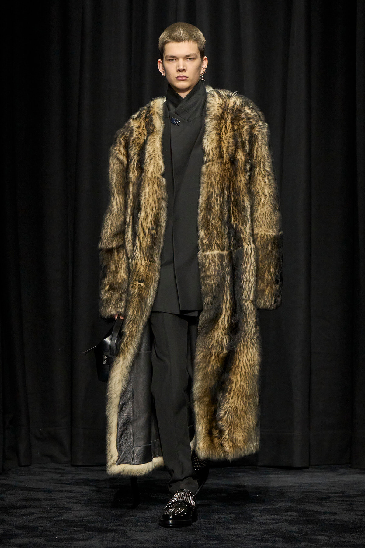 Model on a runway wearing a long fur coat over a black outfit, carrying a black bag, against dark curtains.