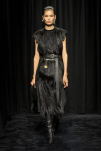 Model walking a runway in a black fringed midi dress with a wide leather belt and knee-high studded boots against a black backdrop.