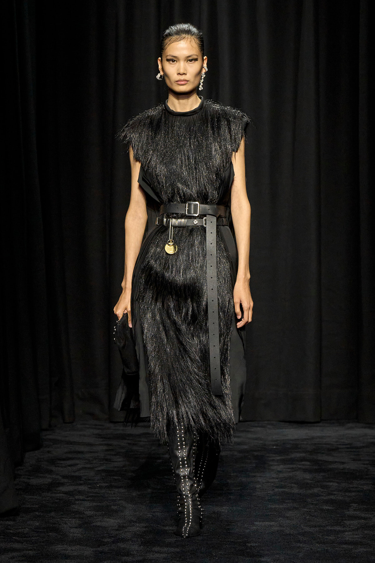 Model walking a runway in a black fringed midi dress with a wide leather belt and knee-high studded boots against a black backdrop.