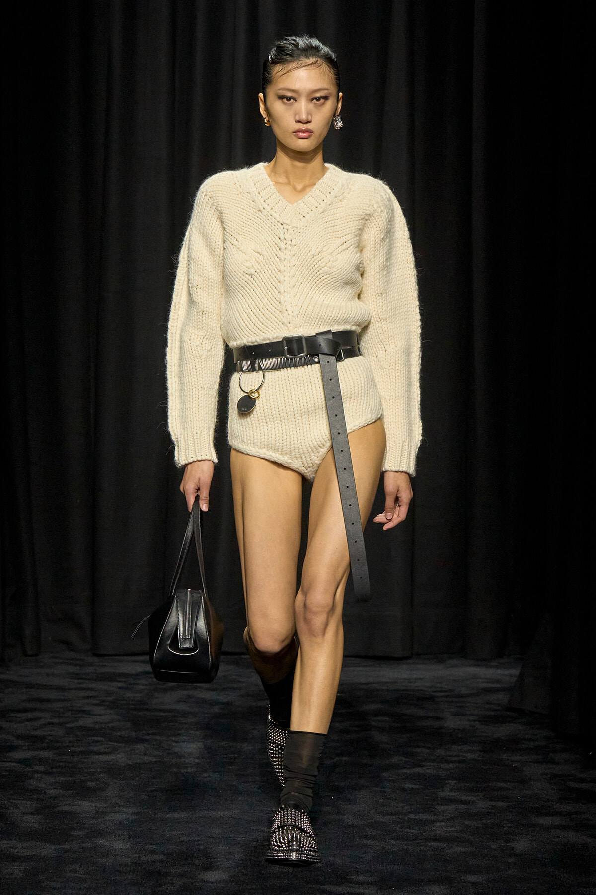 Model walks a runway in a chunky cream knit sweater with matching knit shorts, cinched with a wide black belt and carrying a small black handbag.