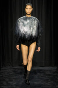 Model walks a runway in a metallic fringe top fading from silver to black, paired with black shorts and ankle boots on a dark stage auras.