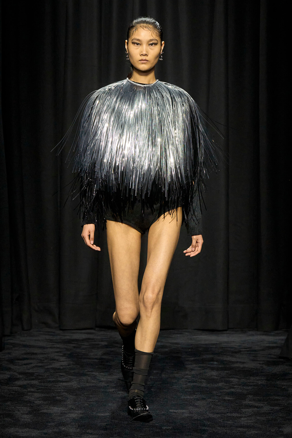 Model walks a runway in a metallic fringe top fading from silver to black, paired with black shorts and ankle boots on a dark stage auras.