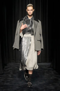 Model on a runway wearing a gray oversized coat over a metallic pleated dress, cinched with a black belt and black studded shoes.