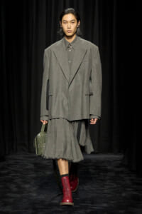 Model on a runway in an oversized gray suit with a pleated, frayed hem skirt, red lace-up boots, and a green studded handbag widening at the side.