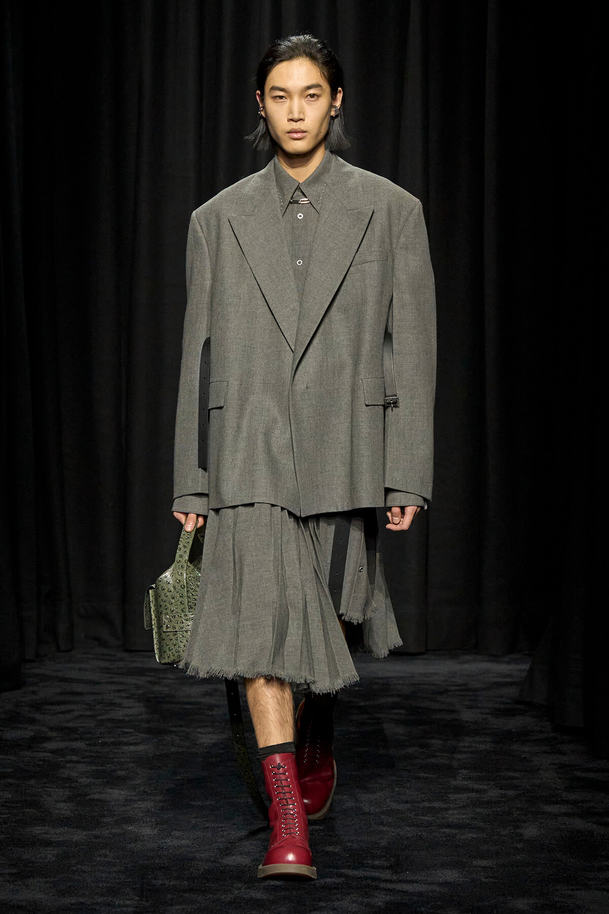 Model on a runway in an oversized gray suit with a pleated, frayed hem skirt, red lace-up boots, and a green studded handbag widening at the side.