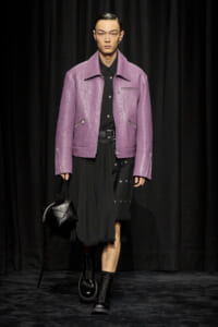 Male model on a runway wearing a lavender textured jacket, black shirt, and a long black pleated skirt, carrying a black geometric handbag and wearing black boots.