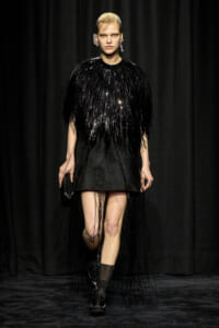 Model walking the runway in a black fringe dress with long tassels and matching accessories, black boots and a small clutch.