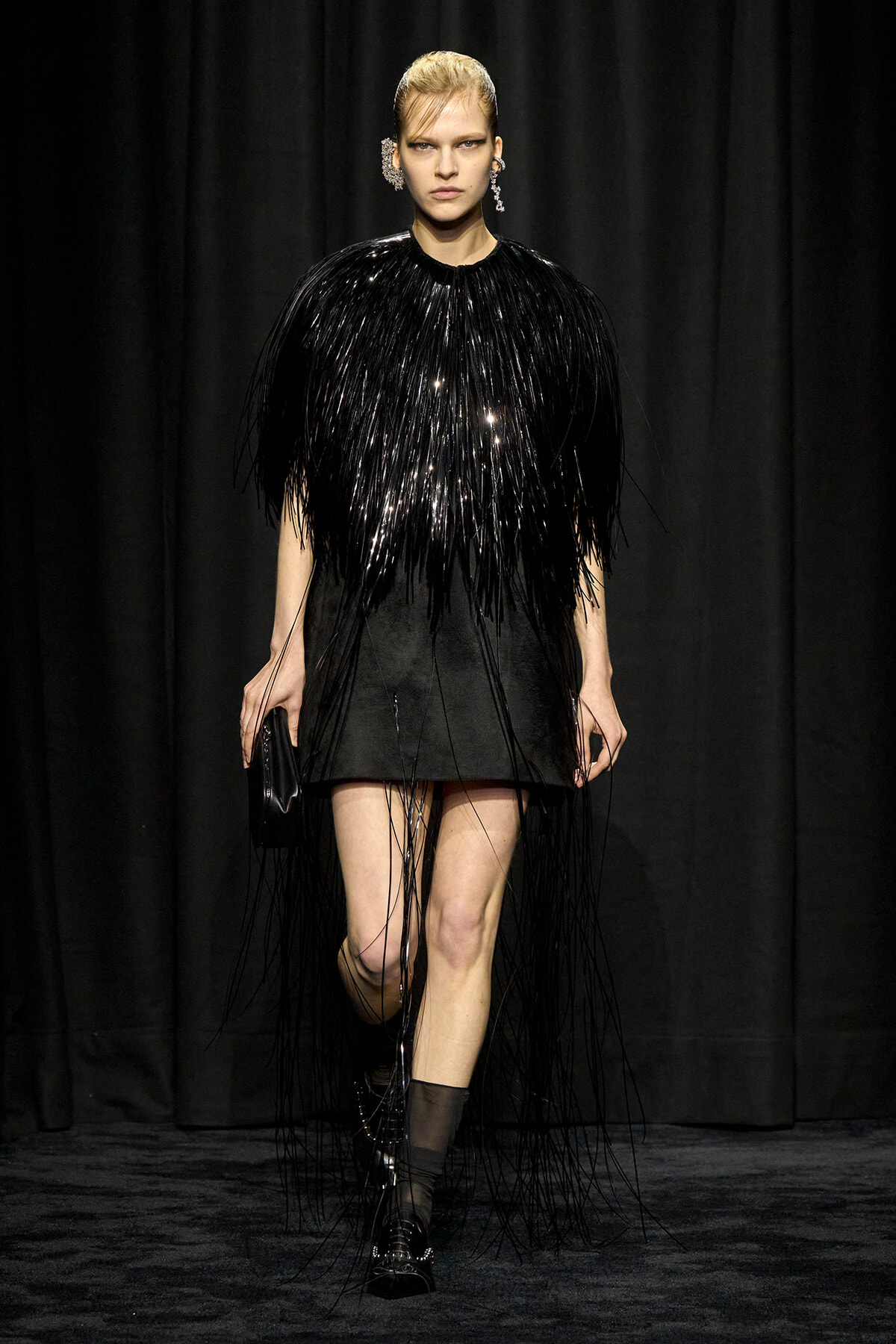 Model walking the runway in a black fringe dress with long tassels and matching accessories, black boots and a small clutch.