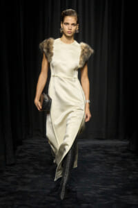 Model on a runway wearing a cream satin dress with fur shoulder accents, large earrings, and a small black clutch.