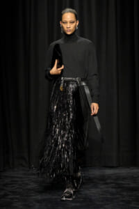 Model walking a runway in a black turtleneck and a long black fringed skirt, holding a black clutch and wearing studded shoes with a wide belt.