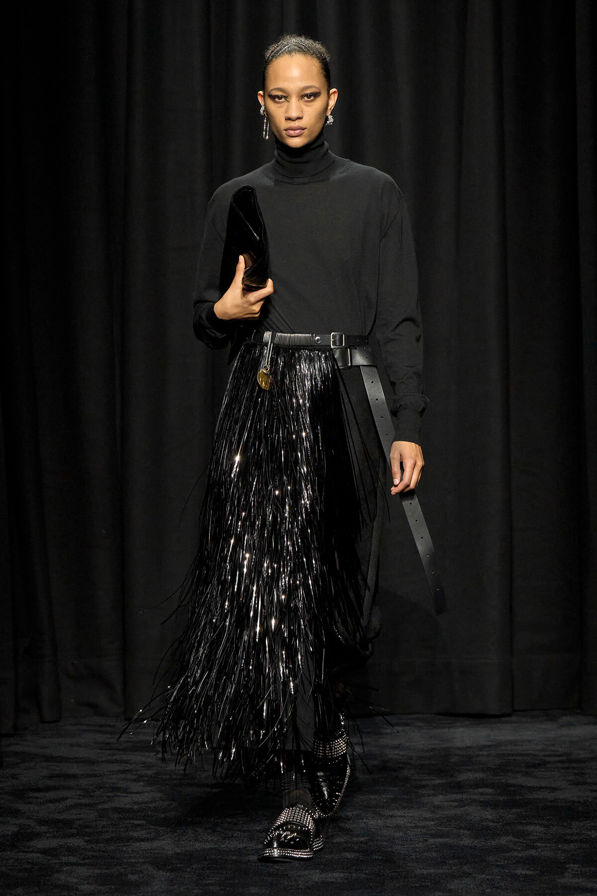 Model walking a runway in a black turtleneck and a long black fringed skirt, holding a black clutch and wearing studded shoes with a wide belt.