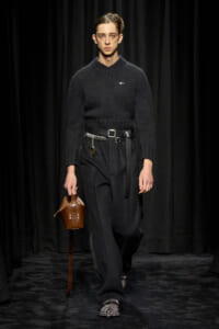 Male model on a runway in a black chunky knit sweater, wide black pants, and a brown perforated basket bag.