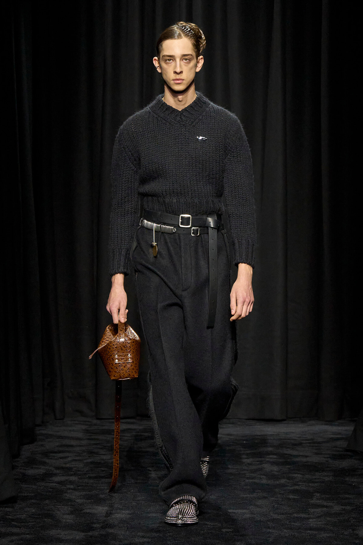 Male model on a runway in a black chunky knit sweater, wide black pants, and a brown perforated basket bag.