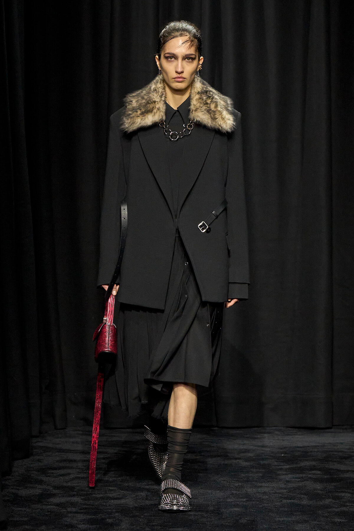 Model walking a runway in a black coat with a fur collar, layered black outfit, and a chunky necklace, carrying a red handbag with a long strap.