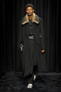 Runway look: a man in a black, fur-collared coat with a layered black outfit, a silver belt, and metallic loafers, carrying a small black clutch.