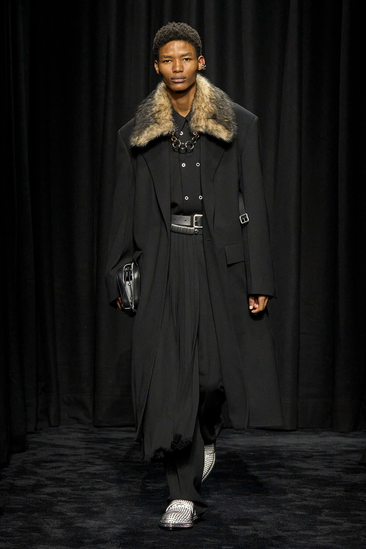 Runway look: a man in a black, fur-collared coat with a layered black outfit, a silver belt, and metallic loafers, carrying a small black clutch.