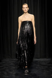 Model walks the runway in a strapless black fringe gown that shimmers under stage lights, with slicked-back hair and statement earrings