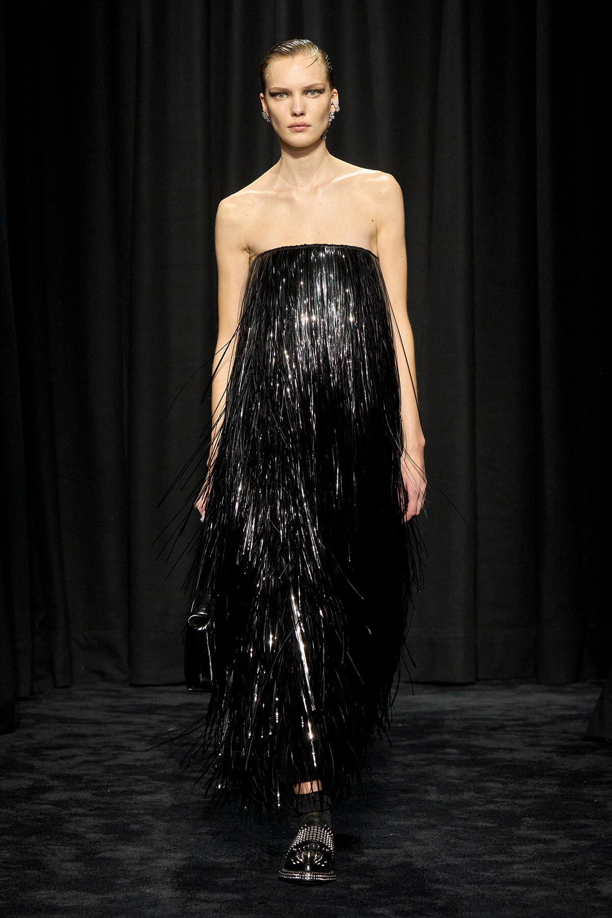 Model walks the runway in a strapless black fringe gown that shimmers under stage lights, with slicked-back hair and statement earrings