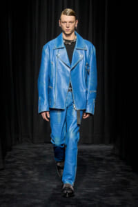 Male model walks the runway wearing a bright blue leather oversized biker jacket and matching pants with silver zippers and studs, black top underneath.
