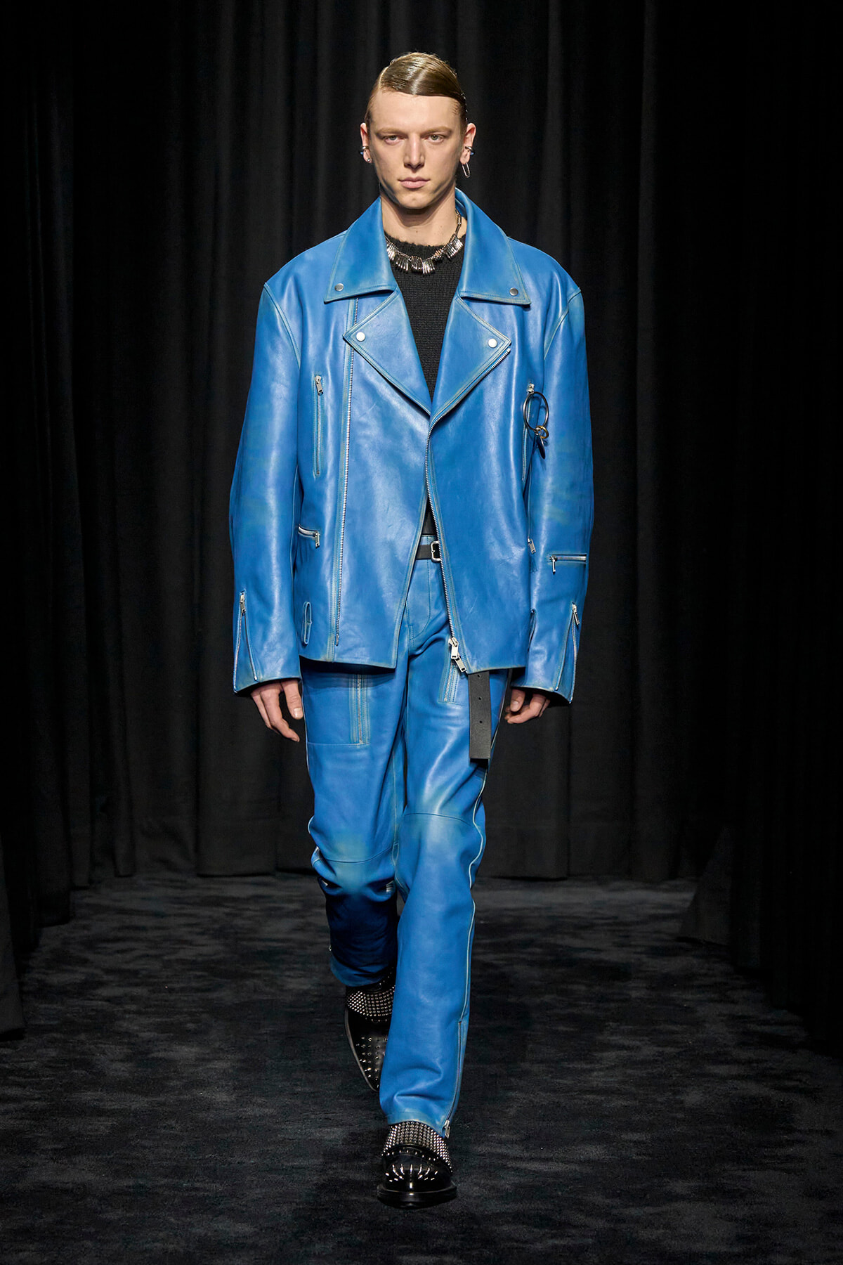 Male model walks the runway wearing a bright blue leather oversized biker jacket and matching pants with silver zippers and studs, black top underneath.