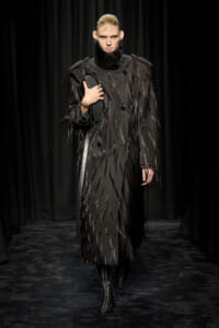 Model on a runway wearing a long black coat with glossy fringe details, holding a dark clutch under her arm.
