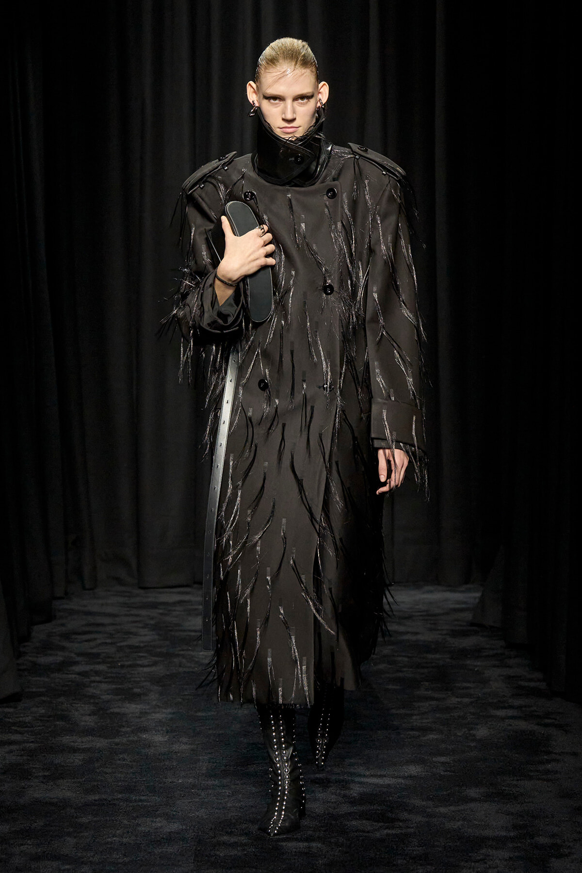 Model on a runway wearing a long black coat with glossy fringe details, holding a dark clutch under her arm.