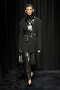 Female model walks a runway in an all-black ensemble: oversized coat with a glossy high-neck panel, studded leather pants, a wide belt, and a black structured handbag.