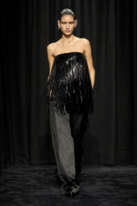 Model walking a runway in a strapless black fringe top and two-tone pants against a black backdrop, mid-stride.