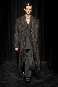 Male runway model wearing a long black fringed coat with metallic strands over a dark suit, black loafers, against a dark backdrop.