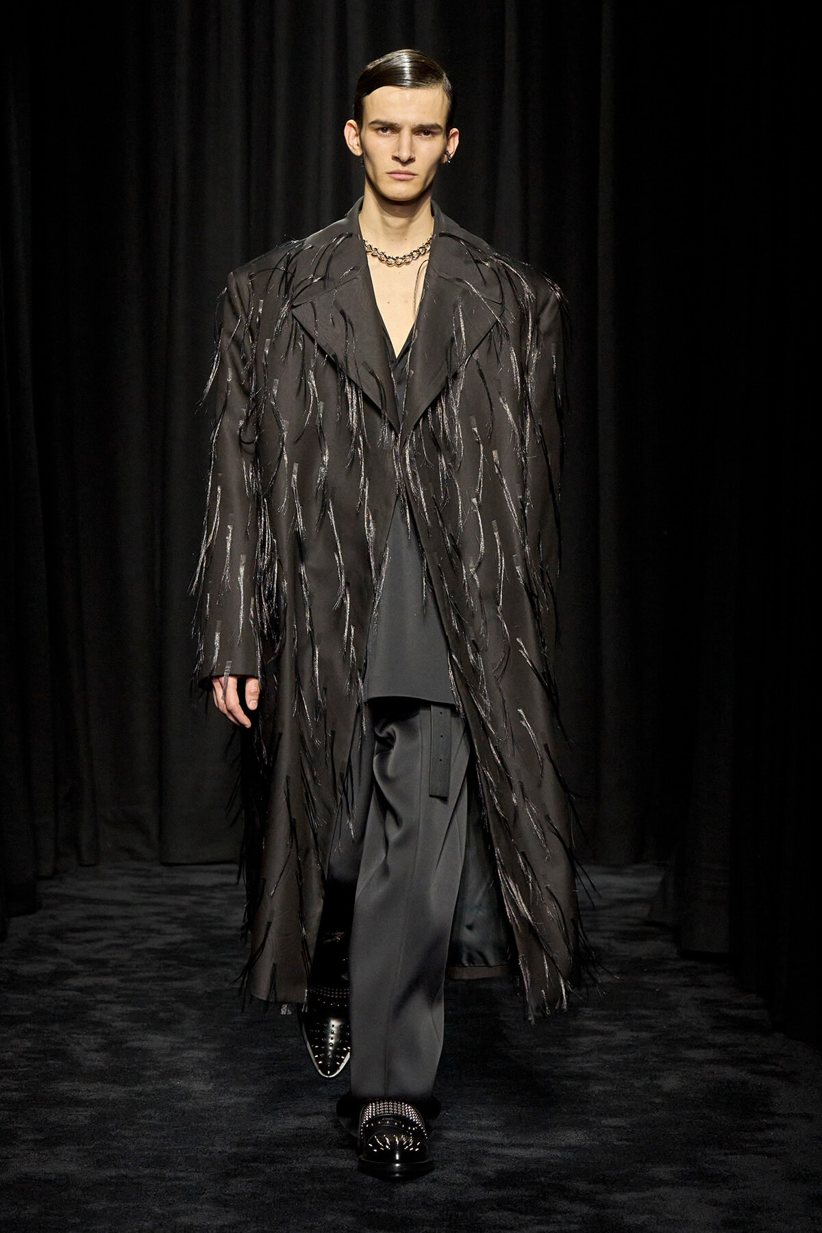 Male runway model wearing a long black fringed coat with metallic strands over a dark suit, black loafers, against a dark backdrop.