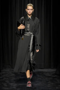 Model on a runway wearing a long black trench with an oversized glossy collar, belt at the waist, and a black clutch bag.