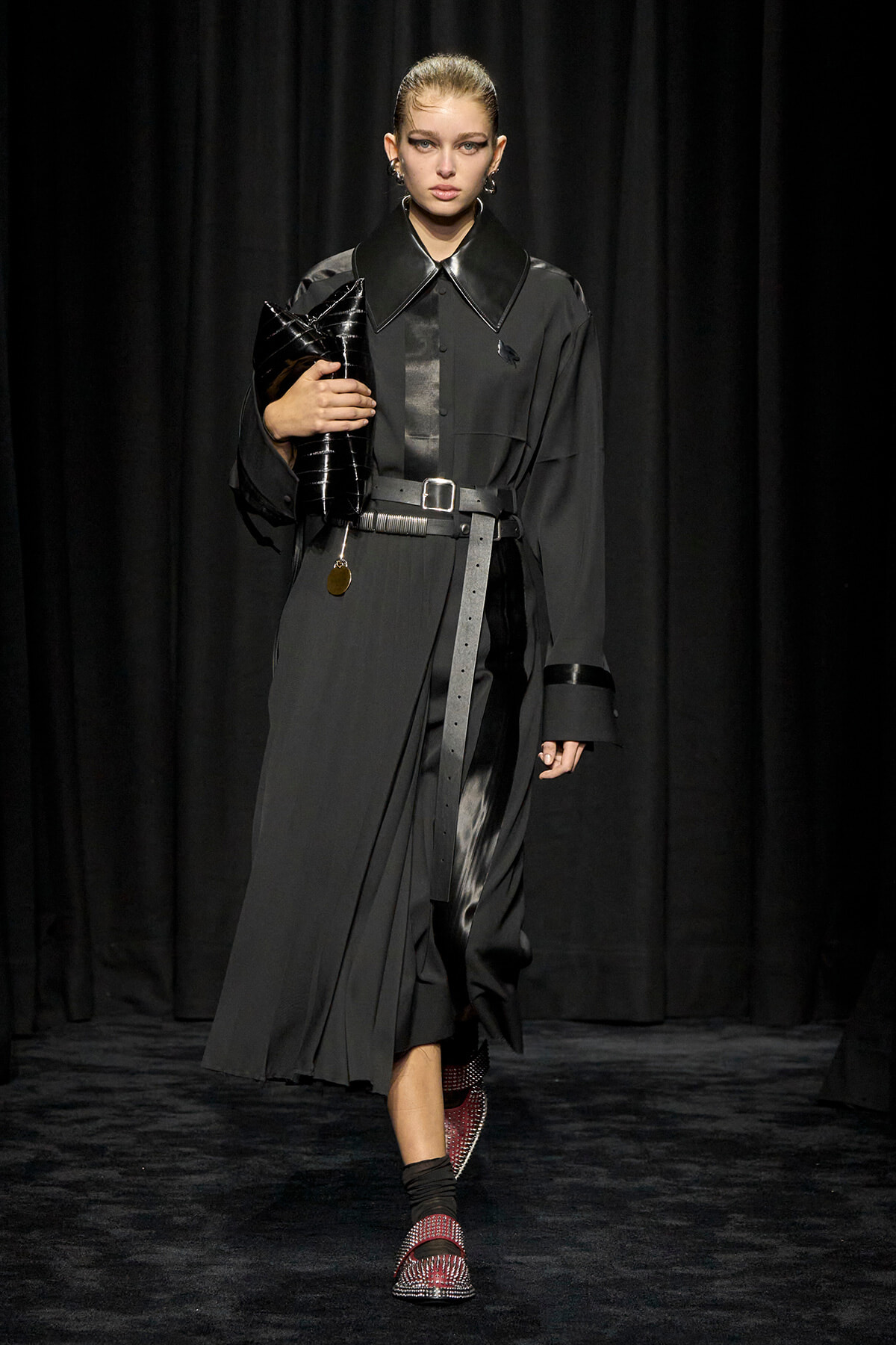 Model on a runway wearing a long black trench with an oversized glossy collar, belt at the waist, and a black clutch bag.