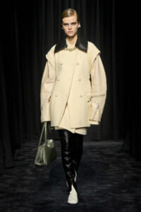 Model on a runway wearing a cream oversized double-breasted coat with black leather lapels, black boots, and a textured green handbag.