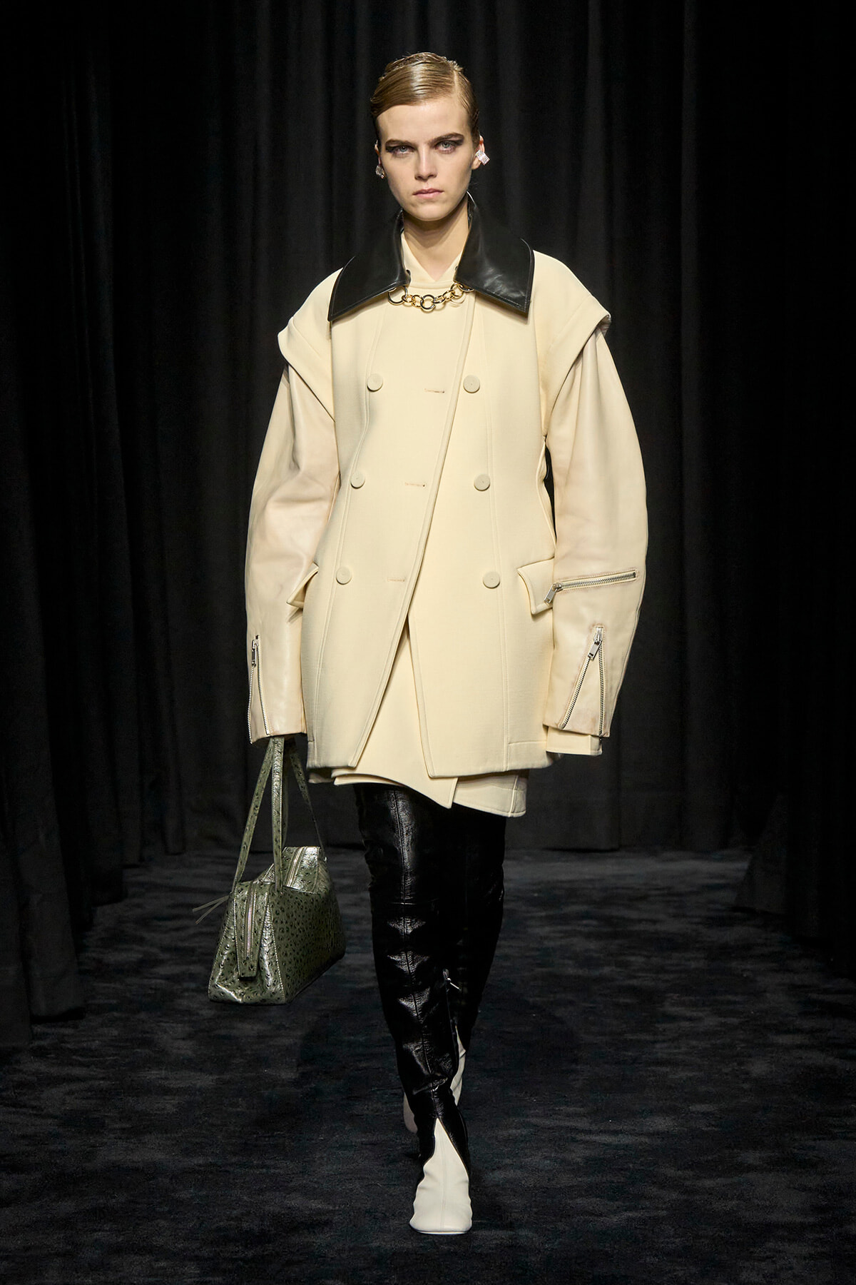Model on a runway wearing a cream oversized double-breasted coat with black leather lapels, black boots, and a textured green handbag.