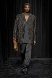 Runway model wearing a dark fringe-embellished blazer over a charcoal outfit, holding a tan clutch.