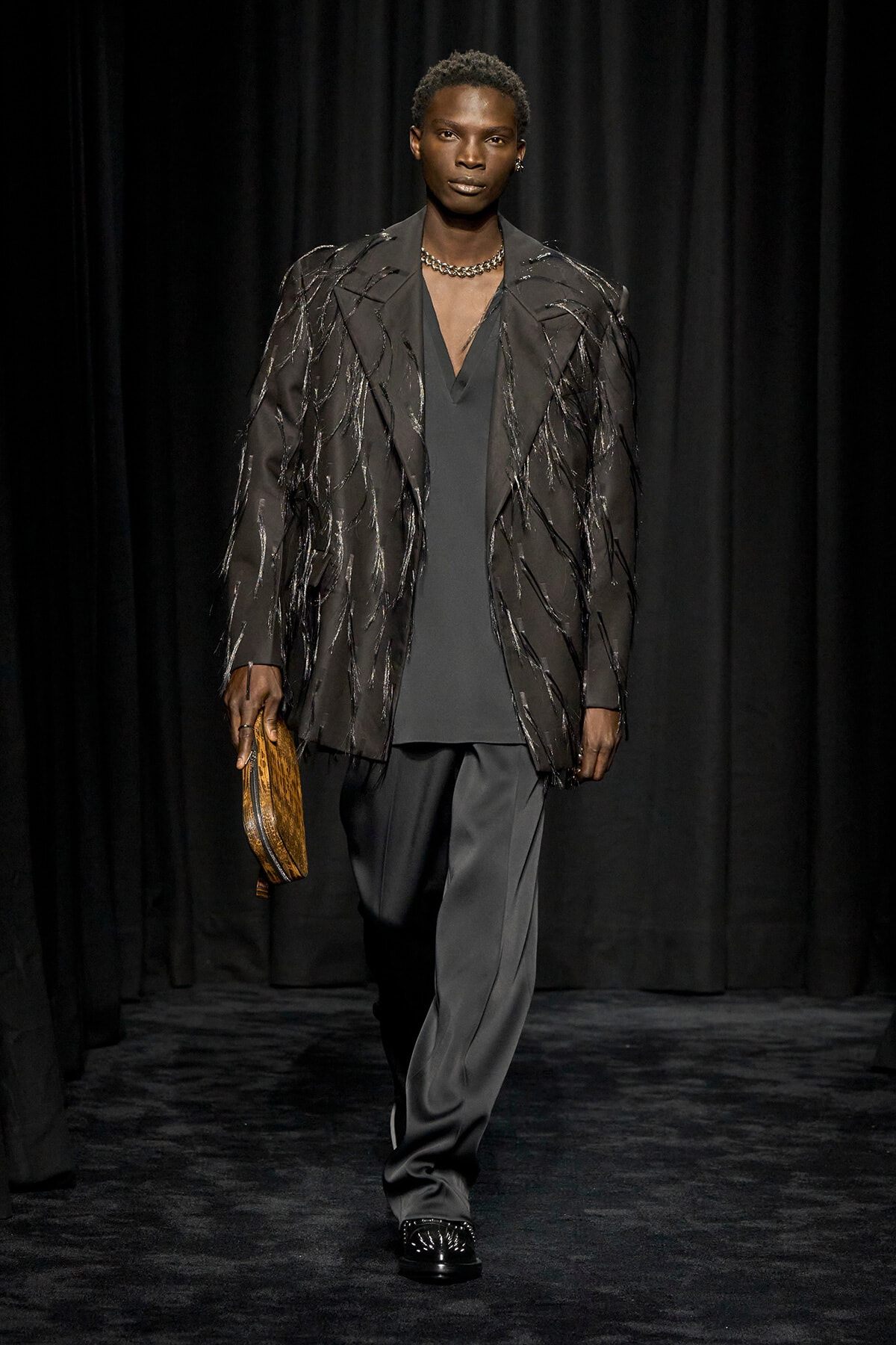 Runway model wearing a dark fringe-embellished blazer over a charcoal outfit, holding a tan clutch.