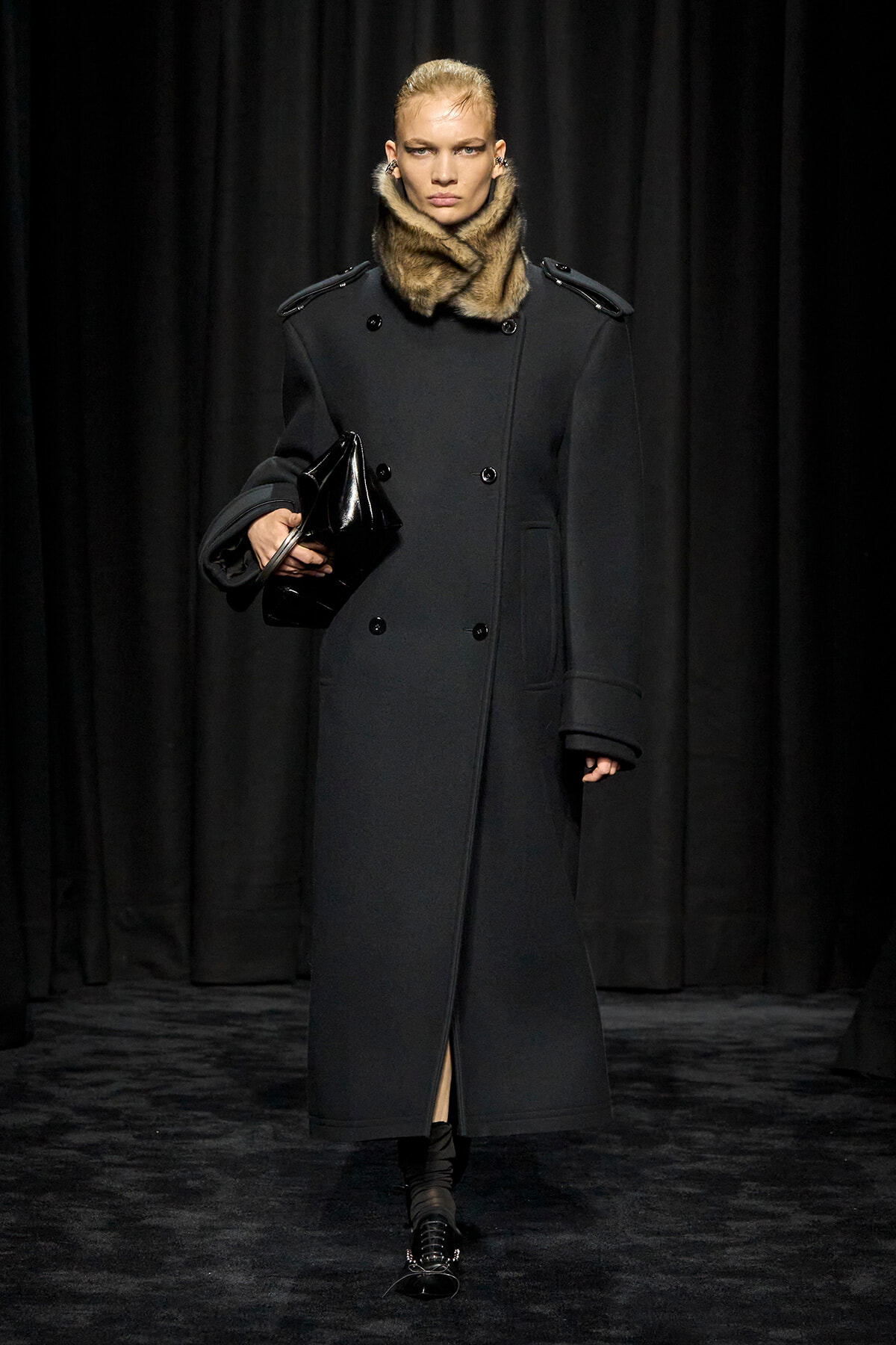 Model on a runway wearing a long black double-breasted coat with a fur collar, carrying a glossy black clutch.
