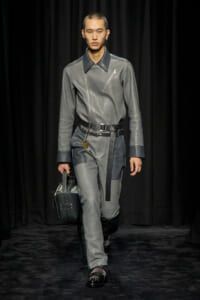 Male model walking a fashion show in a gray leather jumpsuit with a black belt, carrying a gray handbag against a dark backdrop.