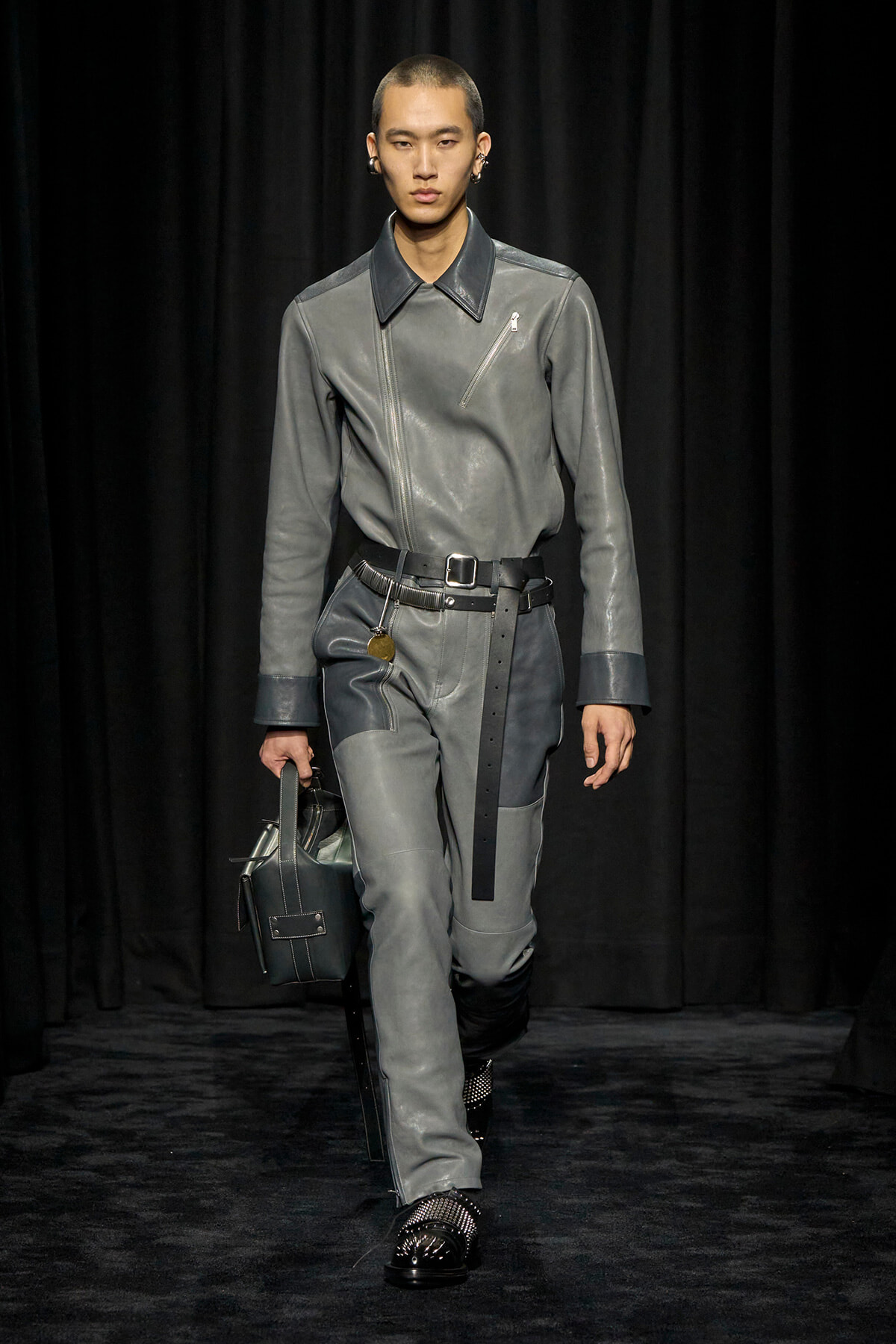 Male model walking a fashion show in a gray leather jumpsuit with a black belt, carrying a gray handbag against a dark backdrop.