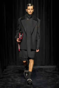 Male model walks the runway in an oversized black coat with wide lapels, black turtleneck, and black footwear, carrying a red clutch.