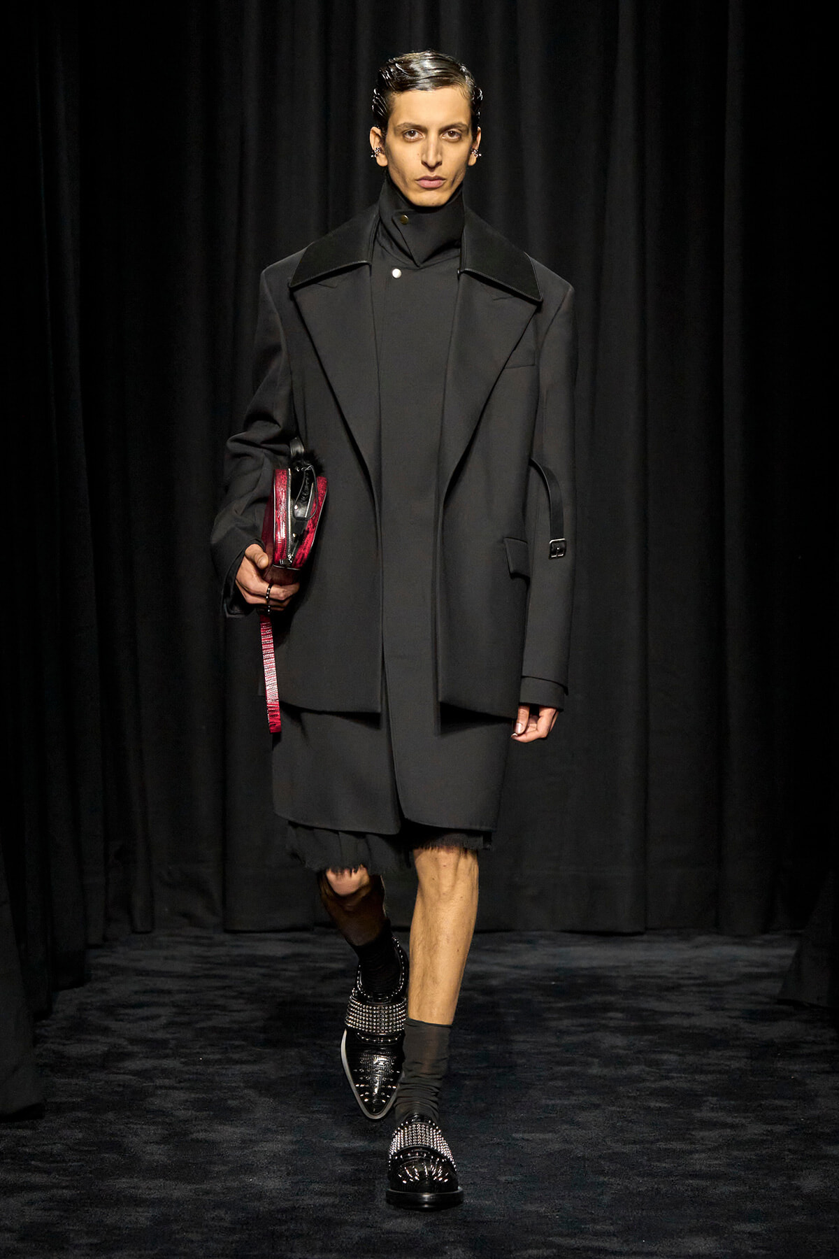Male model walks the runway in an oversized black coat with wide lapels, black turtleneck, and black footwear, carrying a red clutch.