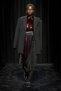 Fashion model walking the runway in a black fringed blazer over a burgundy sequin top and pleated skirt, with a wide belt and studded shoes.